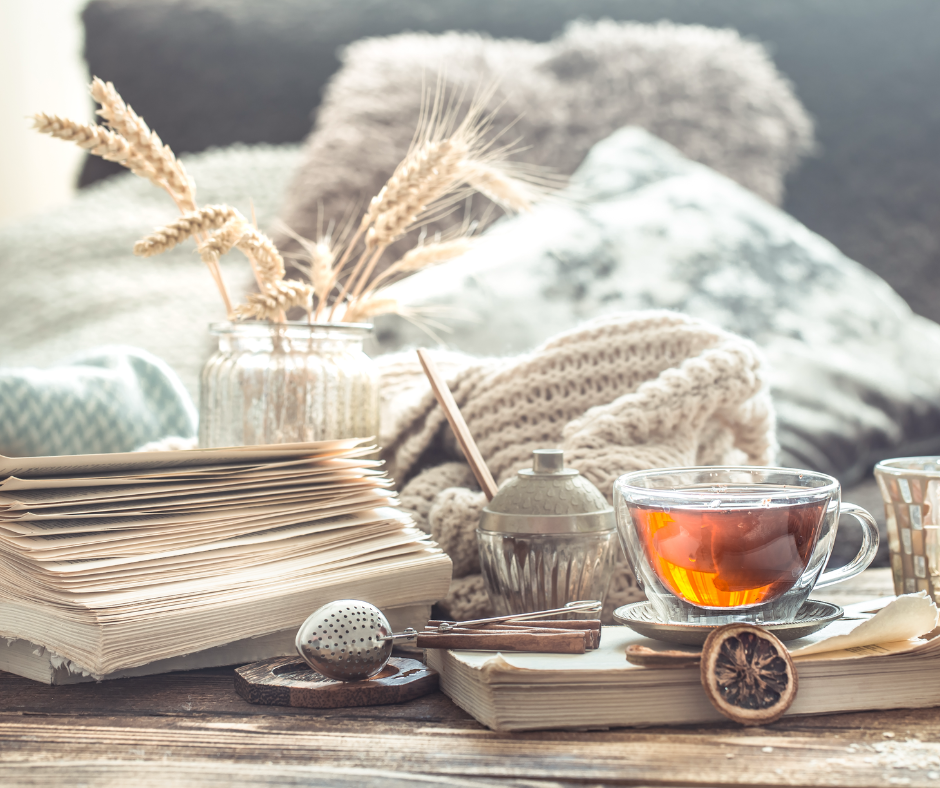 Image shows the simple meaning of hygge. simple and natural. relaxed nordic setting with an open book, tea cup, vase with dried flowers and soft furnishings