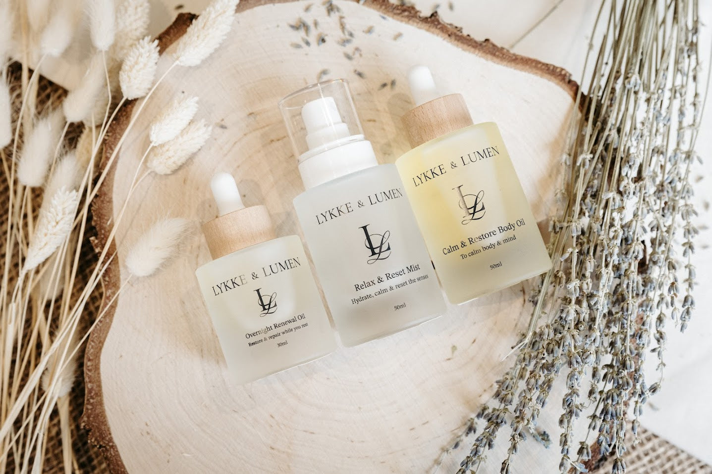 Lykke & Lumen Overnight Renewal Oil, Relax & Reset Mist and Calm & Restore Body Oil arranged on a wood slice with dried lavender and pampas grass — natural botanical skincare for menopause