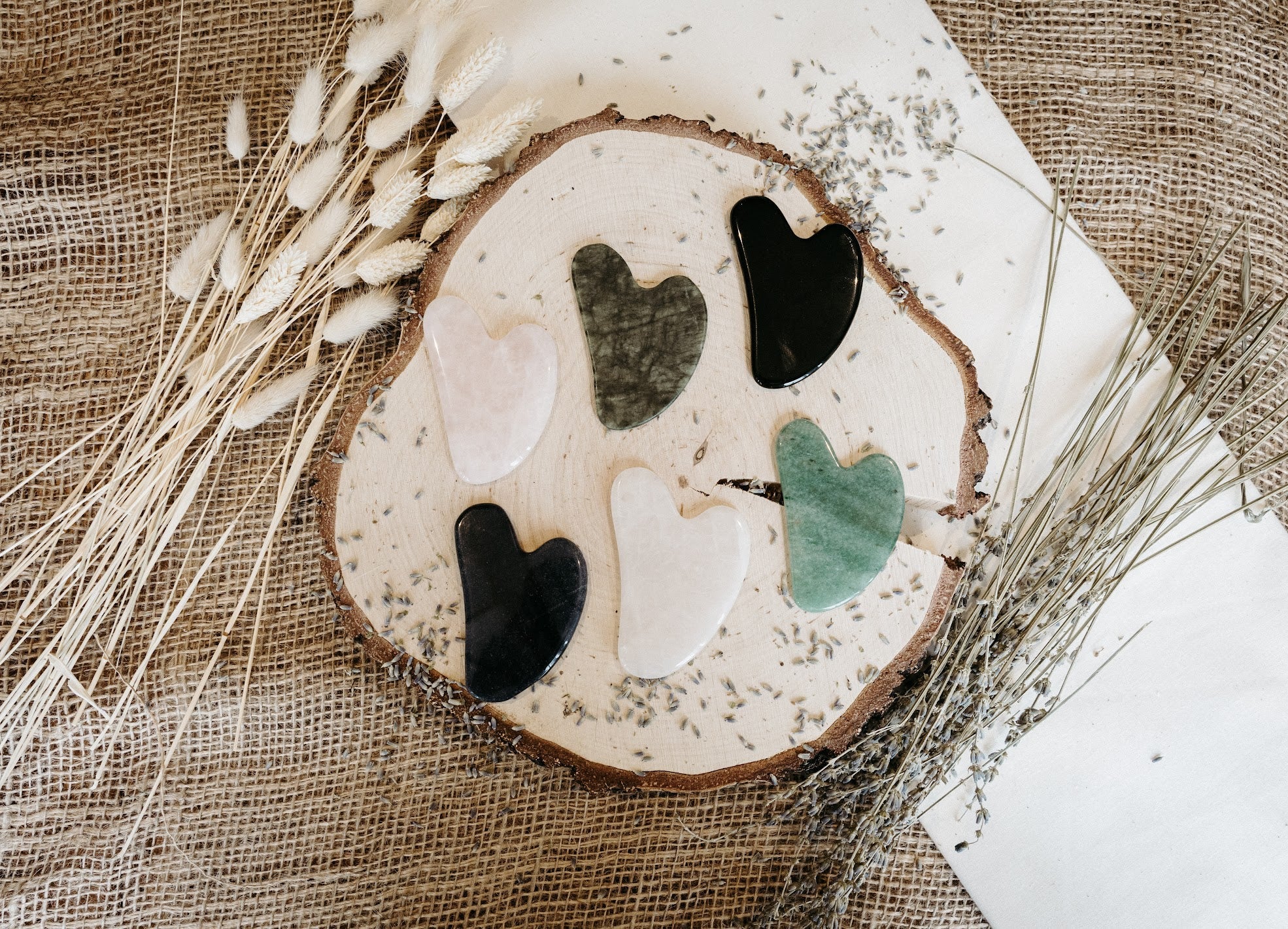 Six Gua-sha gemstones on a wooden slice, surrounded by dried plants on a textured surface.