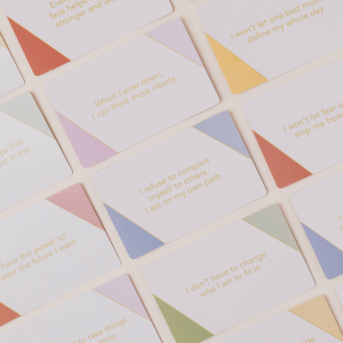 A close-up, top-down view of a set of square cards with rounded corners, featuring inspirational quotes printed in gold text over pastel geometric triangle backgrounds in orange, yellow, purple, blue, green, and pink.