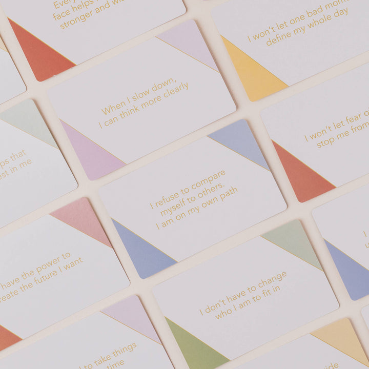A close-up, top-down view of a set of square cards with rounded corners, featuring inspirational quotes printed in gold text over pastel geometric triangle backgrounds in orange, yellow, purple, blue, green, and pink.