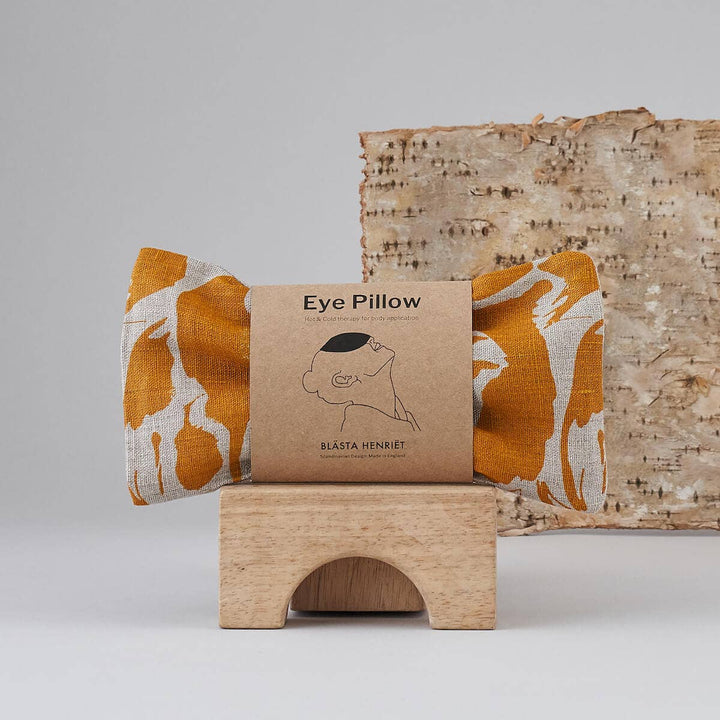 Eye pillow with brown packaging featuring a brand logo on a wooden stand against a neutral background