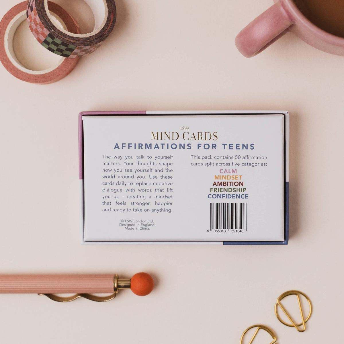 A boxed card deck labeled 'Mind Cards Affirmations for Teens' lies on a pale pink surface, surrounded by styled desk props including washi tape, a pink mug, a wooden pen, and gold rings.