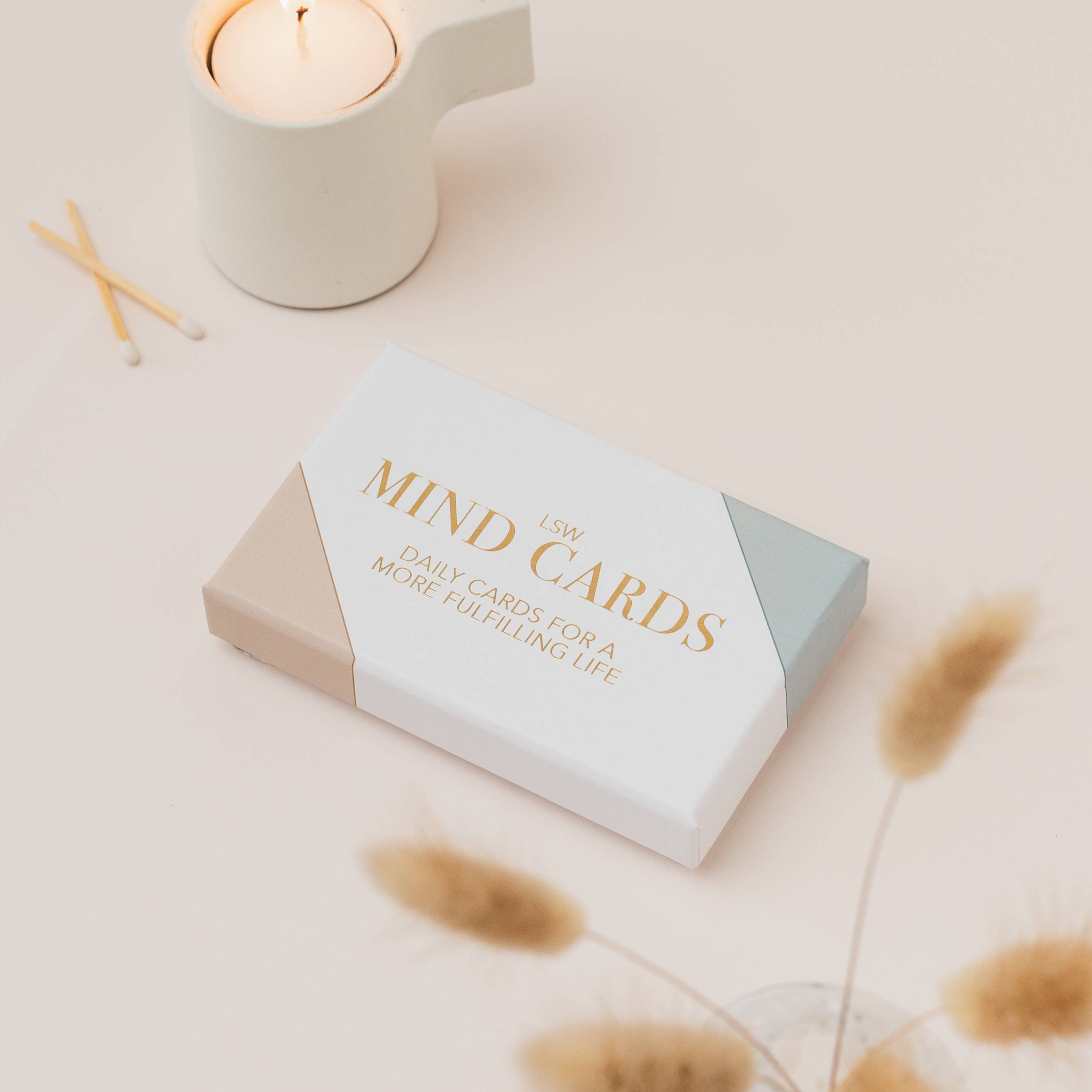 Box of 'MIND GARDS' daily cards for a more fulfilling life on a light surface with a candle and dried plants.