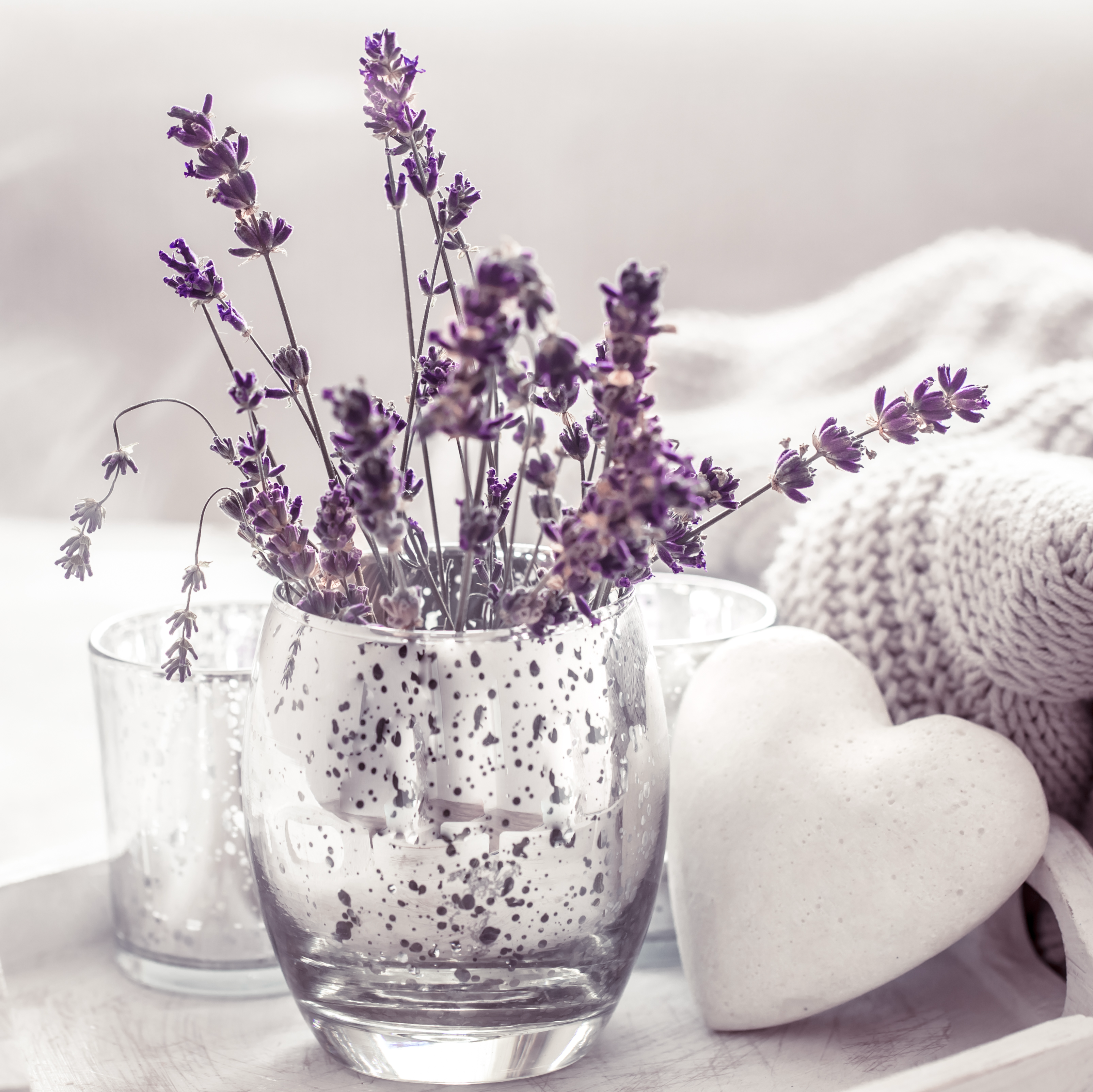 Glass with lavender flowers on a white surface with a heart-shaped object and textured fabric in the background.