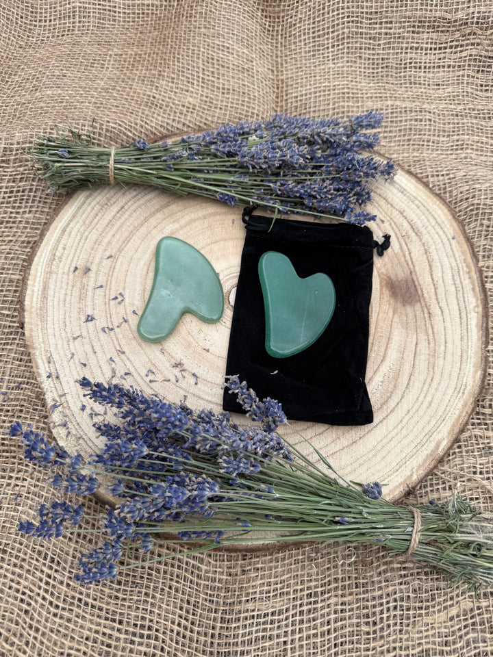 Two green jade Gua-sha face massage gemstones and a black velvet pouch, styled on a round wood slice with lavender bundles on a burlap background.