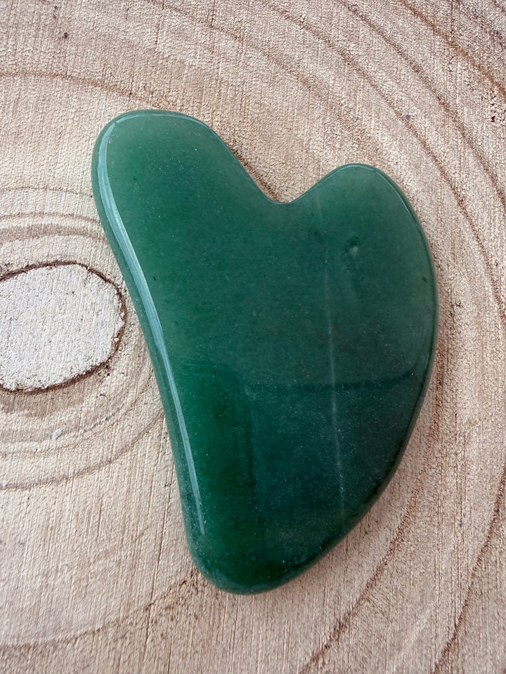 A green jade gua sha resting on a textured wood slice background.