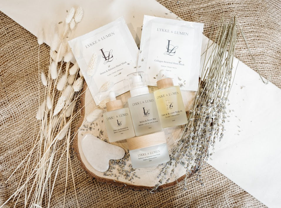 Lykke & Lumen botanical skincare and wellness collection with facial oil, calming mist, body oil, face polish, sheet masks and a gua sha arranged with dried botanicals.