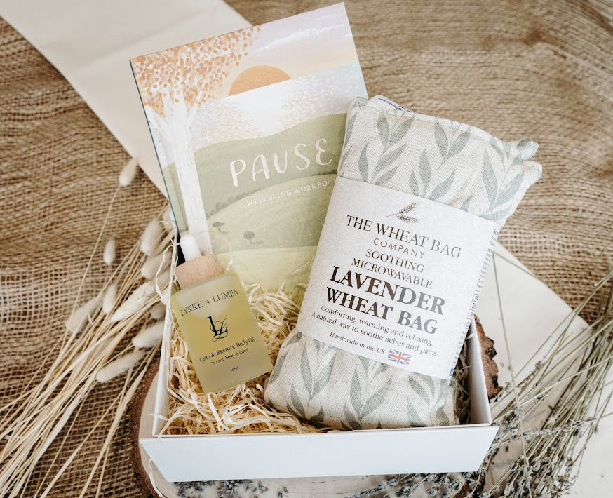 Lykke & Lumen wellness gift box featuring Calm & Restore Body Oil, a lavender wheat bag and wellbeing workbook styled in a luxury self-care setting with natural textures and dried botanicals.