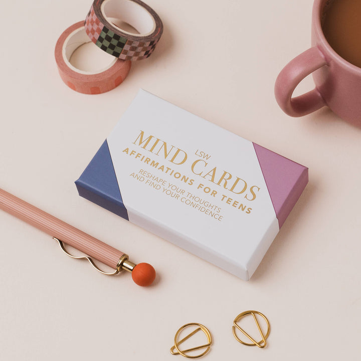 A boxed set of affirmation cards for teens lies on a light beige surface, styled with a pink pen, washi tape rolls, a pink mug, and two gold ring binder clips. The box is white with blue and lavender geometric blocks and gold text reading 'MIND CARDS AFFIRMATIONS FOR TEENS' 