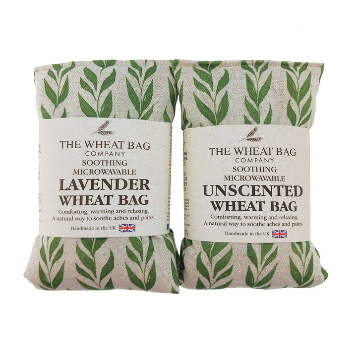 Two packaged soothing wheat bags from The Wheat Bag Company on a white background. One bag is lavender and the other is unscented, both wrapped in a beige linen fabric with a green leaf pattern and a white paper label.