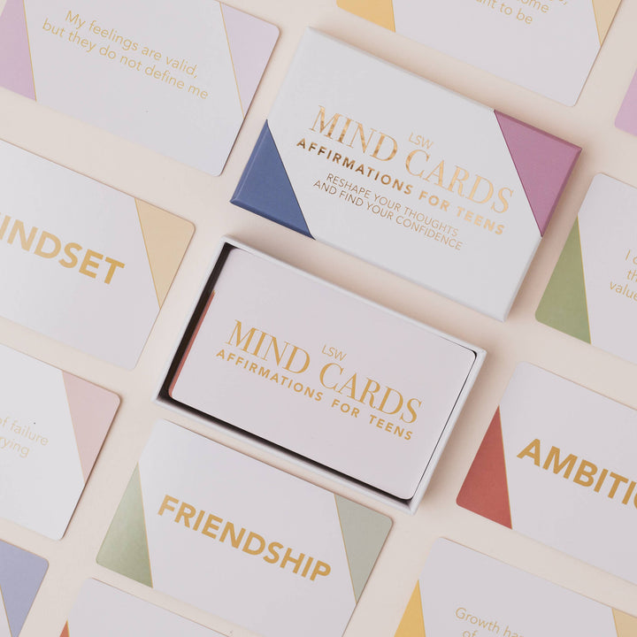 A boxed set of affirmation cards for teens arranged on a light cream background. The product includes a white box labeled 'LSW MIND CARDS AFFIRMATIONS FOR TEENS' and several cards with pastel geometric corner accents and gold text, including cards reading 'MINDSET,' 'FRIENDSHIP,' and 'AMBITION.