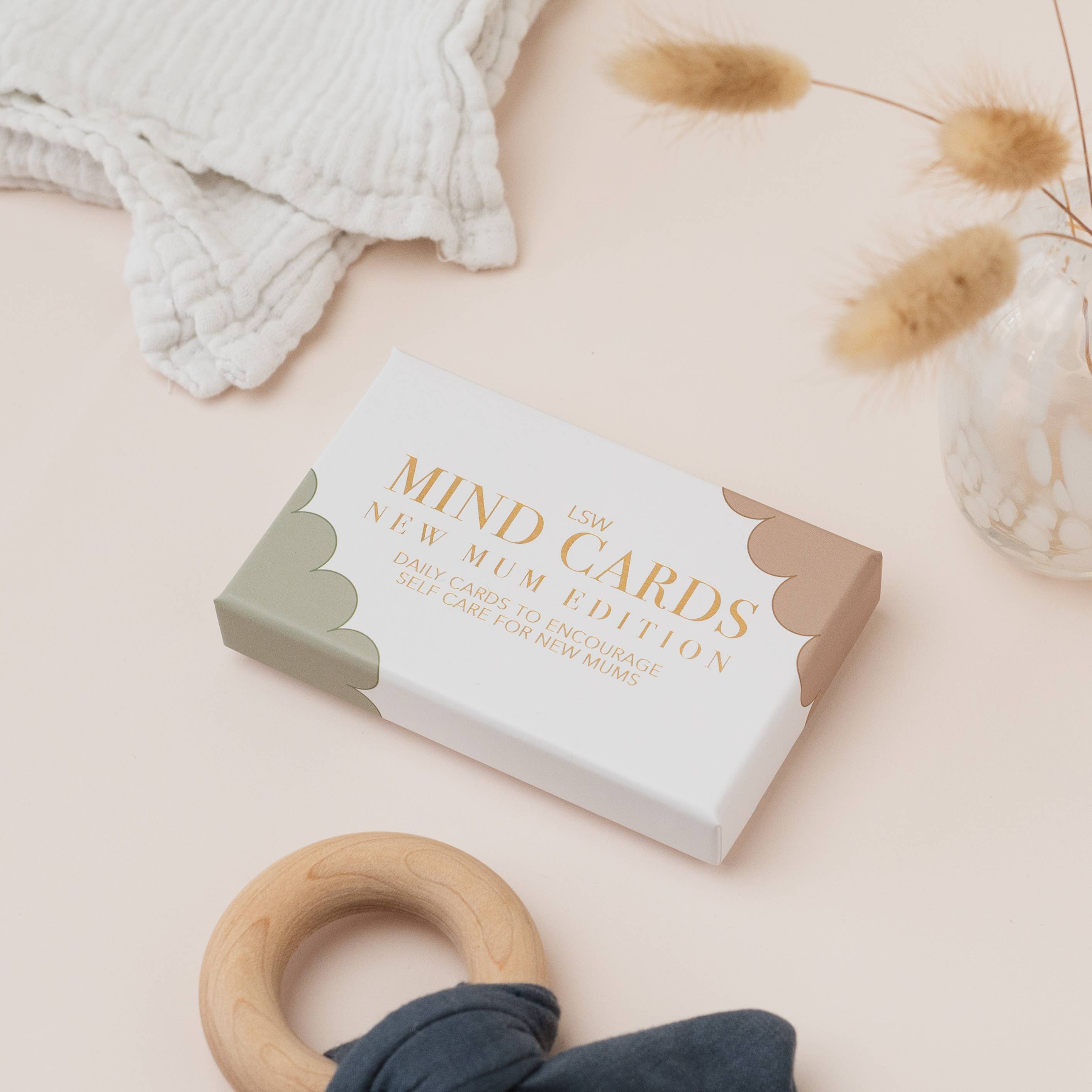 Box of 'New Mum Edition' cards on a light pink surface with a wooden ring and fabric.